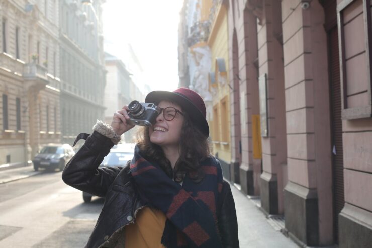 A woman holding a video camera, wearing a trilby hat. Depiction of big hats, and their versatility with plethora of outfits.